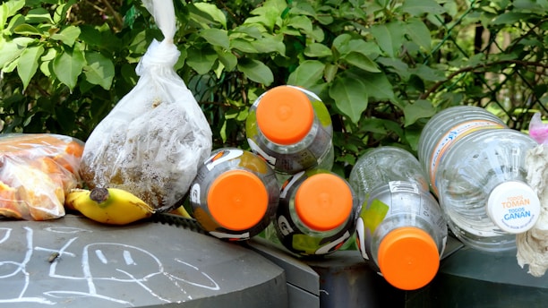 Various discarded items including plastic bottles with orange caps, a banana, and bags containing what appears to be organic waste are placed on top of a graffiti-covered surface with green foliage in the background.