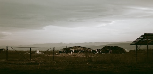 A rural landscape with a cloudy sky. A group of cows is gathered around a barn-like structure surrounded by open fields. Wooden fences and a partially constructed or dismantled shed are visible. The background features distant mountains under a broad, expansive sky.