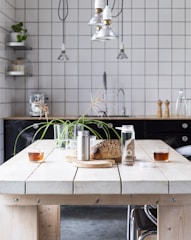 A modern kitchen setting with a light wooden table featuring two glasses of tea, a loaf of bread on a cutting board, a metal container, and a glass bottle with a cork lid. The background includes a black countertop, a tiled backsplash, a metal faucet, pepper mills, and hanging pendant lights. A potted plant is placed on the table, and there are industrial-style shelves with various items.