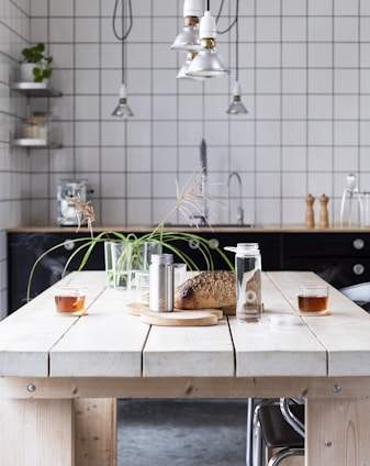 A modern kitchen setting with a light wooden table featuring two glasses of tea, a loaf of bread on a cutting board, a metal container, and a glass bottle with a cork lid. The background includes a black countertop, a tiled backsplash, a metal faucet, pepper mills, and hanging pendant lights. A potted plant is placed on the table, and there are industrial-style shelves with various items.