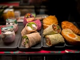 An assortment of food items is displayed on trays. There are several types of wraps filled with vegetables, chicken, and other ingredients, arranged neatly. Behind these are croissants and a pastry with an egg in the center. Additionally, there are two cups with granola and yogurt topped with blueberries.