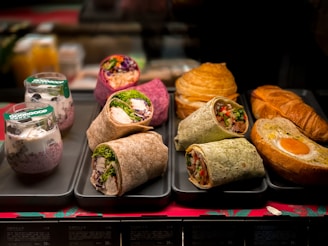 An assortment of food items is displayed on trays. There are several types of wraps filled with vegetables, chicken, and other ingredients, arranged neatly. Behind these are croissants and a pastry with an egg in the center. Additionally, there are two cups with granola and yogurt topped with blueberries.