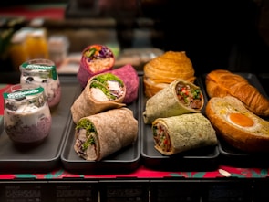 An assortment of food items is displayed on trays. There are several types of wraps filled with vegetables, chicken, and other ingredients, arranged neatly. Behind these are croissants and a pastry with an egg in the center. Additionally, there are two cups with granola and yogurt topped with blueberries.