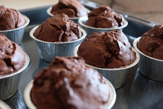 Several chocolate muffins are placed in individual metallic baking cups on a tray. The muffins have a rich, brown color and appear freshly baked with a slightly cracked top, indicating a moist interior.
