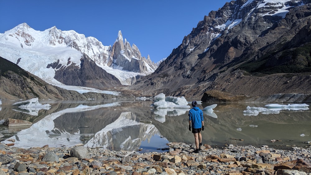 person in blue jacket and blue denim jeans standing on rocky ground near lake and snow