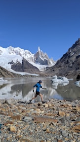 man in blue shirt and black shorts standing on rocky shore near snow covered mountain during