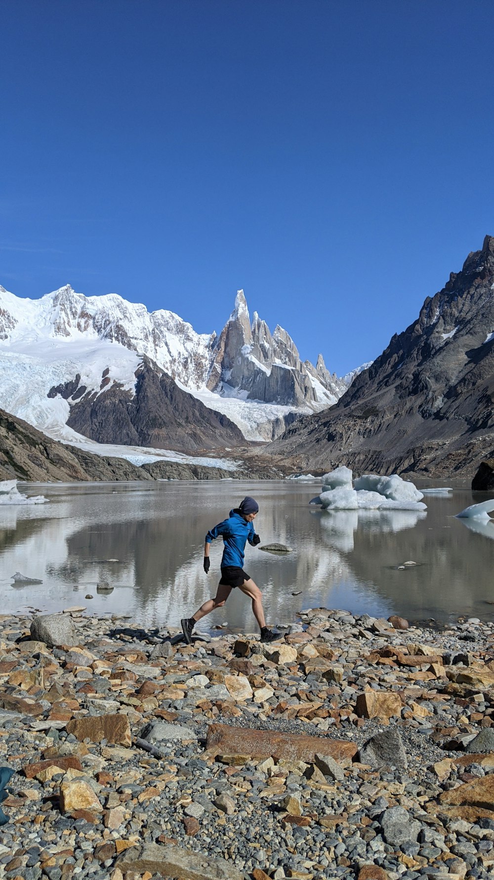 man in blue shirt and black shorts standing on rocky shore near snow covered mountain during