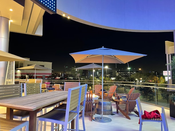 A modern outdoor patio setting at night, featuring a wooden table with high chairs and several wooden lounge chairs arranged around circular tables. Large umbrellas provide shade, and the area is illuminated by overhead lights, creating a cozy ambiance. The background includes city lights and a partially visible building.