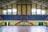 An indoor sports arena with a basketball court. The arena has blue, tiered seating sections with some red accents, and large, yellow walls with windows allowing natural light. The court flooring is visible in the foreground, and a basketball hoop with a blue backboard is centered. There is a symmetrical layout with staircases leading to the seating area.