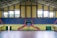 An indoor sports arena with a basketball court. The arena has blue, tiered seating sections with some red accents, and large, yellow walls with windows allowing natural light. The court flooring is visible in the foreground, and a basketball hoop with a blue backboard is centered. There is a symmetrical layout with staircases leading to the seating area.