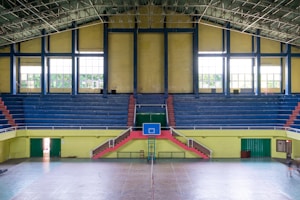 An indoor sports arena with a basketball court. The arena has blue, tiered seating sections with some red accents, and large, yellow walls with windows allowing natural light. The court flooring is visible in the foreground, and a basketball hoop with a blue backboard is centered. There is a symmetrical layout with staircases leading to the seating area.