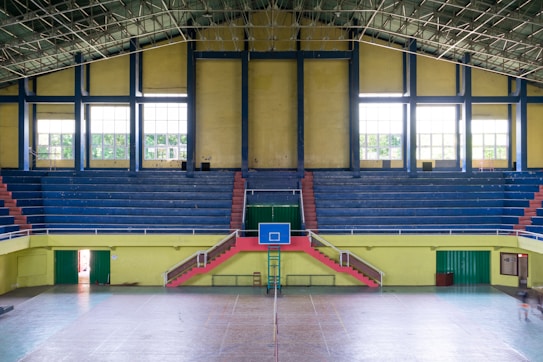 An indoor sports arena with a basketball court. The arena has blue, tiered seating sections with some red accents, and large, yellow walls with windows allowing natural light. The court flooring is visible in the foreground, and a basketball hoop with a blue backboard is centered. There is a symmetrical layout with staircases leading to the seating area.