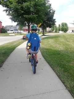 A child wearing a helmet and blue backpack is riding a bicycle down a suburban sidewalk. The pathway is surrounded by grass, and there are residential houses and trees in the background under a cloudy sky.
