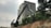 A large excavator is positioned on a dirt slope next to an urban construction site, with a tall residential building in the background. A worker wearing a safety helmet stands near the excavator. The scene is surrounded by lush greenery, and the sky is overcast.