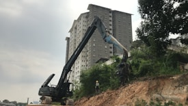 A large excavator is positioned on a dirt slope next to an urban construction site, with a tall residential building in the background. A worker wearing a safety helmet stands near the excavator. The scene is surrounded by lush greenery, and the sky is overcast.