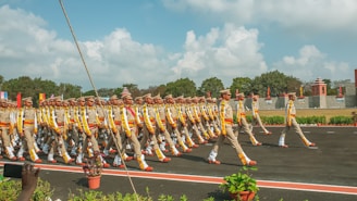 A group of uniformed individuals is marching in formation on a road. They are wearing beige outfits with colorful accents and matching hats. The setting appears to be an outdoor event with a backdrop of trees, a wall, and blue skies with scattered clouds. Potted plants and a red strip line the pathway.