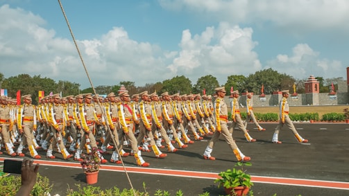 A group of uniformed individuals is marching in formation on a road. They are wearing beige outfits with colorful accents and matching hats. The setting appears to be an outdoor event with a backdrop of trees, a wall, and blue skies with scattered clouds. Potted plants and a red strip line the pathway.