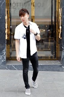 A young man stands on a city street wearing a white baseball-style shirt with text, black jeans, and checkered sneakers. He holds sunglasses in one hand and wears a wristwatch. The background features a modern building entrance with reflective glass and golden accents.