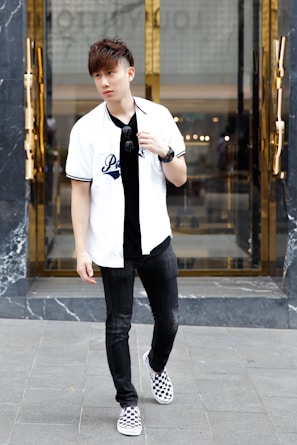 A young man stands on a city street wearing a white baseball-style shirt with text, black jeans, and checkered sneakers. He holds sunglasses in one hand and wears a wristwatch. The background features a modern building entrance with reflective glass and golden accents.