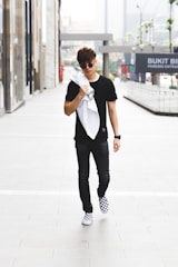 A young man in casual attire walks confidently down an urban sidewalk. He wears a black shirt, dark pants, and checkered sneakers, and carries a white jacket or towel over his shoulder. The background features glass-fronted buildings and signage indicating a shopping area.