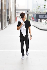 A young man in casual attire walks confidently down an urban sidewalk. He wears a black shirt, dark pants, and checkered sneakers, and carries a white jacket or towel over his shoulder. The background features glass-fronted buildings and signage indicating a shopping area.