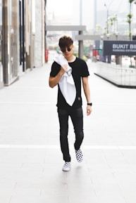 A young man in casual attire walks confidently down an urban sidewalk. He wears a black shirt, dark pants, and checkered sneakers, and carries a white jacket or towel over his shoulder. The background features glass-fronted buildings and signage indicating a shopping area.