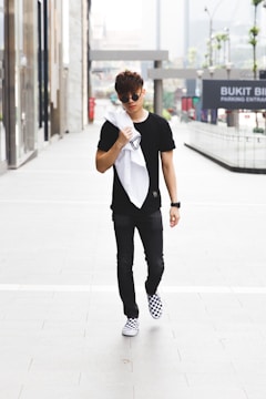 A young man in casual attire walks confidently down an urban sidewalk. He wears a black shirt, dark pants, and checkered sneakers, and carries a white jacket or towel over his shoulder. The background features glass-fronted buildings and signage indicating a shopping area.
