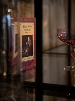 A book titled 'Hans Børlis beste dikt' is displayed with a decorative pink and white cover, featuring a black and white photograph of a man. Next to the book, there is a wine glass with a deep red tint on a reflective glass surface.