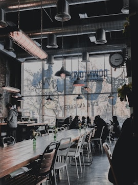 A stylish cafe interior with industrial design elements, featuring metal ductwork and large hanging lamps. The seating area is populated with people sitting at long wooden tables, some working on laptops. Large windows let in natural light and display artwork of a rooster silhouette advertising a bakery. Decorative green bottles with flowers are placed on the tables.