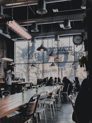 A stylish cafe interior with industrial design elements, featuring metal ductwork and large hanging lamps. The seating area is populated with people sitting at long wooden tables, some working on laptops. Large windows let in natural light and display artwork of a rooster silhouette advertising a bakery. Decorative green bottles with flowers are placed on the tables.