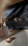 A black leather handbag with a gold clasp and strap is prominently displayed. Next to it is a faceted glass perfume bottle with a gold top, and a pair of gold and pearl earrings is laid out in front.