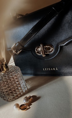 A black leather handbag with a gold clasp and strap is prominently displayed. Next to it is a faceted glass perfume bottle with a gold top, and a pair of gold and pearl earrings is laid out in front.