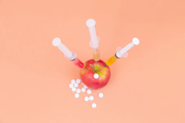 A red apple with three syringes inserted into it, surrounded by white tablets on a peach-colored background.