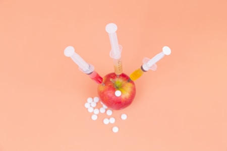 A red apple with three syringes inserted into it, surrounded by white tablets on a peach-colored background.