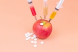 A red apple with three syringes inserted into it, each containing different colored liquids. White pills are scattered around the base of the apple on a peach-colored background.