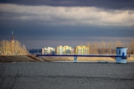 A body of water in the foreground, bordered by a concrete dam, with a blue structure and railing extending over it. There is a backdrop of several beige and yellow apartment buildings surrounded by leafless trees under a cloudy, overcast sky.