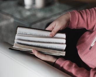 Hands holding an open book, with pages being turned. The person is wearing a pink sweater, and the scene appears to be indoors with a sofa and a table in view.
