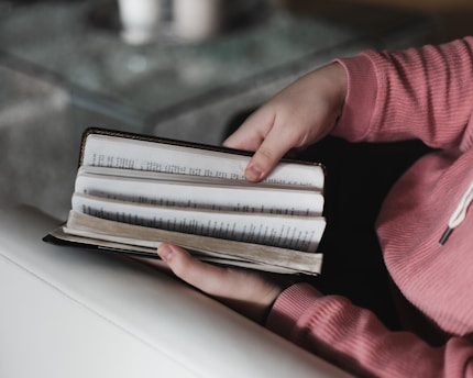 Hands holding an open book, with pages being turned. The person is wearing a pink sweater, and the scene appears to be indoors with a sofa and a table in view.