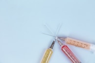 Three syringes with varying colored liquids are arranged on a light blue background. The syringes are positioned with their needles pointing inward, forming a star-like pattern.