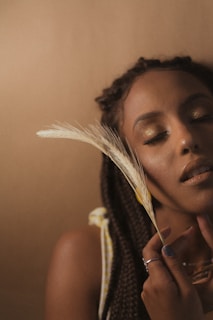 A serene portrait of a woman with her eyes closed, holding a feather near her face. She has braided hair and a peaceful expression, set against a soft, earthy-toned background.