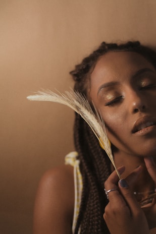 A serene portrait of a woman with her eyes closed, holding a feather near her face. She has braided hair and a peaceful expression, set against a soft, earthy-toned background.