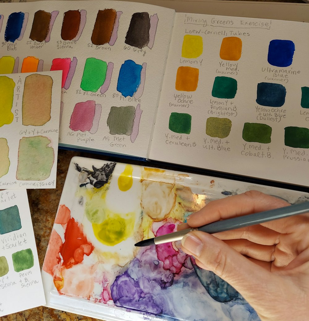 a person's hand holding a pen over a palette of watercolor paints