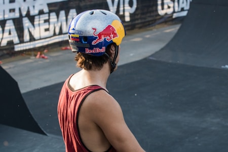 A person wearing a helmet with a vibrant design, including logos and graphics, stands in a skate park. The individual is wearing a sleeveless shirt, gazing towards the ramps and the advertising banners visible in the background.