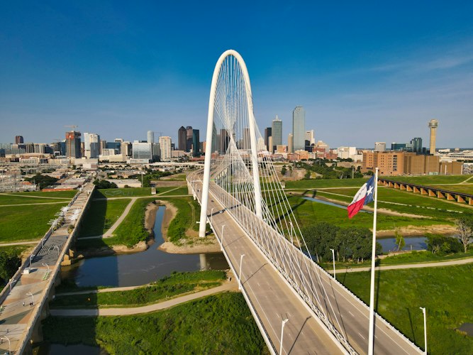 Aerial view of the white arch of the Margaret Hunt Hill Bridge crossing the Trinity River, with the downtown Dallas skyline and Reunion Tower behind it and a Texas flag in the foreground