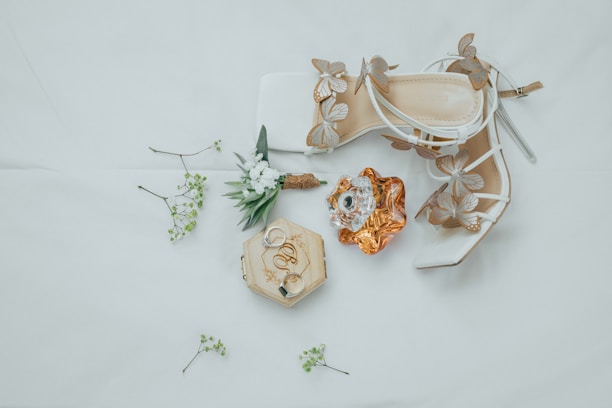 An arrangement of elegant bridal accessories includes a pair of white high-heeled shoes adorned with butterfly designs, a bottle of perfume with an ornate cap, a wooden box with engraved rings, and a small bouquet of greenery and white flowers scattered on a soft, neutral background.