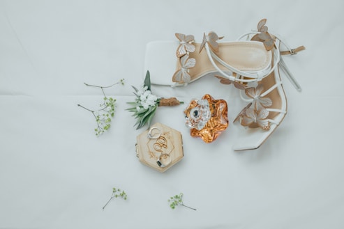 An arrangement of elegant bridal accessories includes a pair of white high-heeled shoes adorned with butterfly designs, a bottle of perfume with an ornate cap, a wooden box with engraved rings, and a small bouquet of greenery and white flowers scattered on a soft, neutral background.