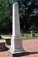 A tall, white stone monument stands on a red brick path, engraved with text commemorating individuals. The monument is surrounded by green grass and trees, with additional grave markers visible in the background.