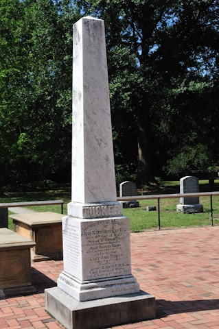 A tall, white stone monument stands on a red brick path, engraved with text commemorating individuals. The monument is surrounded by green grass and trees, with additional grave markers visible in the background.