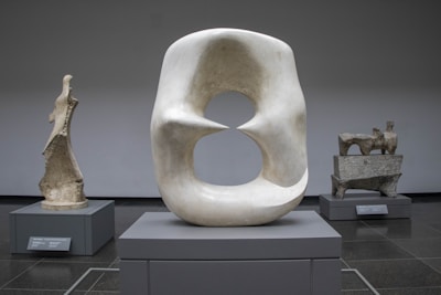 Three abstract stone sculptures are displayed within a gallery setting. The central piece is a large, smooth sculpture with a hollowed center, while the other two are rougher in texture and smaller in size. Each sculpture is placed on a pedestal within a minimalist space with a neutral background.
