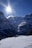 A snowy mountain landscape features a bright sunlight shining prominently in the sky, creating lens flares. The scene includes steep, rugged mountains covered with snow, a small wooden cabin situated on a snow-covered hill in the foreground, and dense evergreen trees. The atmosphere is serene and majestic.